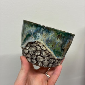 May include: A ceramic bowl featuring a textured lower section with a raised, hand-painted stone wall pattern in shades of gray and brown, topped with a smooth, glossy glaze in vibrant swirls of deep green, blue, and cream colors.