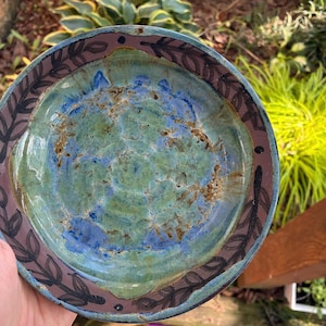 May include: A round ceramic bowl featuring a mottled green and blue glazed interior with a glossy finish. The wide rim is painted with a dark brown, repeating leaf pattern against a muted mauve background, creating a distinct border.
