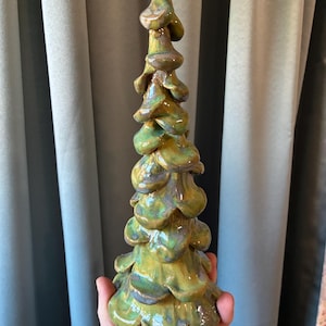 May include: A tall, conical ceramic sculpture shaped like a stylized evergreen tree. The surface features a glossy, multi-tonal glaze with blended shades of moss green, olive, and hints of earthy brown, creating a layered, organic texture throughout.