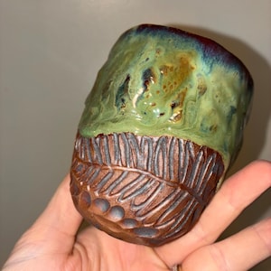 May include: A ceramic cup featuring a two-tone design with a smooth, glossy green and blue glazed upper section and a textured, unglazed brown lower section. The bottom portion displays a carved fern leaf pattern and circular indentations.