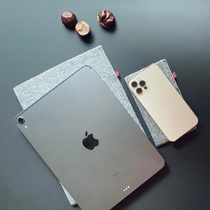 May include: A gray iPad with a white Apple logo on a gray fabric surface. A gold iPhone with a black camera lens is on a gray fabric surface with a red and white stripe.