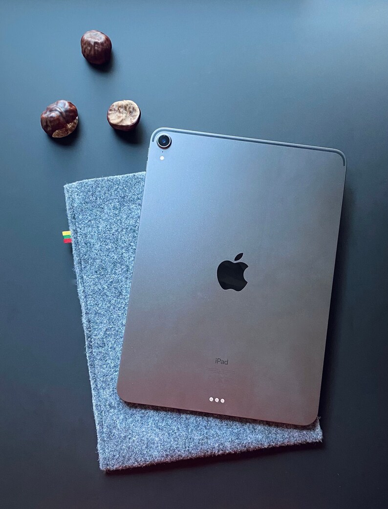 May include: A silver iPad with a black Apple logo and the word "iPad" printed on the back. The iPad is lying on a gray fabric surface.