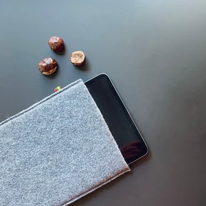 Puede incluir: Una funda de tableta de fieltro gris con una tableta negra en su interior. Tres castañas marrones están esparcidas sobre una superficie gris.