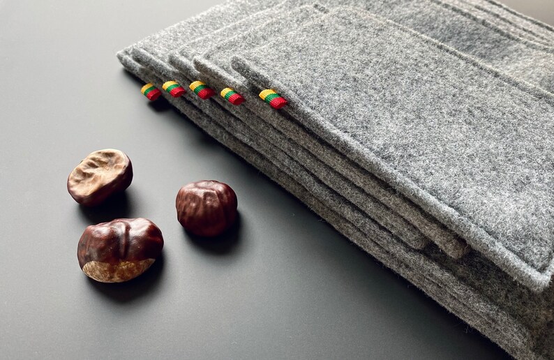 May include: A stack of gray felt coasters with yellow, green, and red ribbon accents. The coasters are arranged on a gray surface with three brown chestnuts.