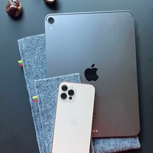 May include: A white iPhone with a black camera lens and a silver iPad with a black Apple logo on a gray surface. The iPad is on top of a gray felt case with yellow and red stripes.