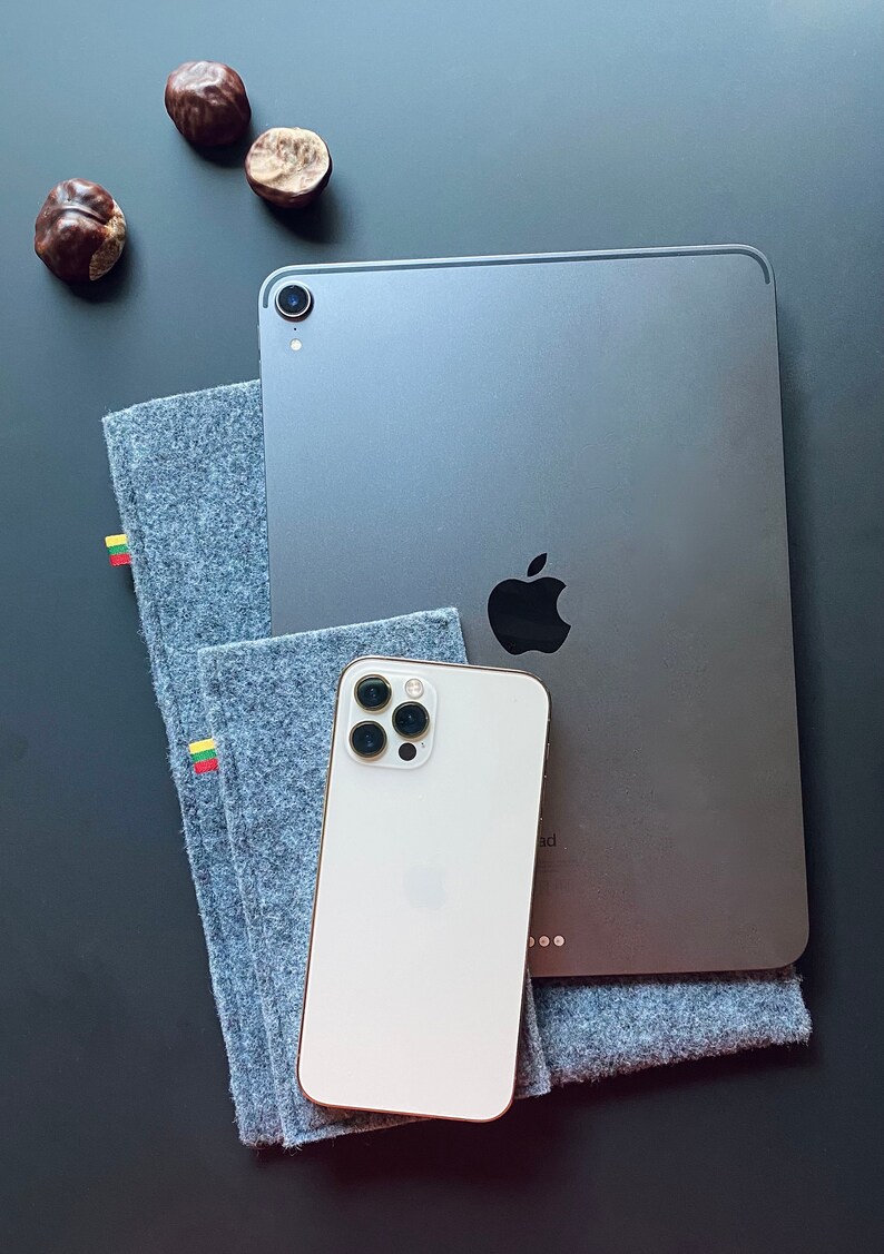 May include: A white iPhone with a black camera lens and a silver iPad with a black Apple logo on a gray surface. The iPad is on top of a gray felt case with yellow and red stripes.