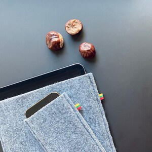 May include: A gray felt tablet sleeve with a small pocket for a phone. The sleeve has a small Lithuanian flag patch on the side. The sleeve is lying on a black surface with three brown chestnuts.