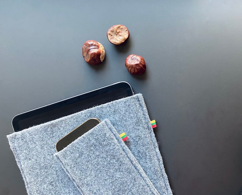 May include: A gray felt tablet sleeve with a small pocket for a phone. The sleeve has a small Lithuanian flag patch on the side. The sleeve is lying on a black surface with three brown chestnuts.