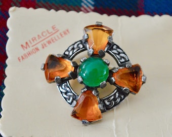 Puede incluir: Un broche de nudo celta de plata con una piedra verde en el centro y cuatro piedras de color ámbar alrededor. El broche está sobre una tarjeta blanca con el texto "Miracle Fashion Jewellery".
