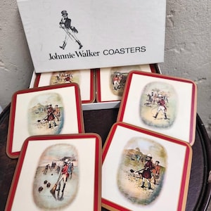 Puede incluir: Un juego de posavasos cuadrados con borde rojo e ilustración vintage. Los posavasos presentan una escena de personas jugando un deporte, y la caja dice "Johnnie Walker COASTERS" con un logotipo de un hombre caminando.
