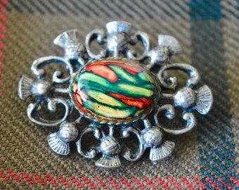 Puede incluir: Un broche de plata con un diseño de esmalte colorido. El diseño presenta un óvalo central con rayas verdes, amarillas y rojas. El broche está rodeado de seis motivos de cardo de plata.