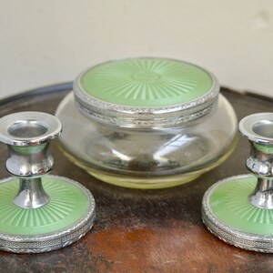 K&ouml;nnte beinhalten: Ein Vintage-Schminktisch-Set mit zwei silbernen Kerzenst&auml;ndern und einem Glasgef&auml;&szlig; mit gr&uuml;nem Deckel. Die Kerzenst&auml;nder haben eine gr&uuml;ne Basis mit einem silbernen Rand.