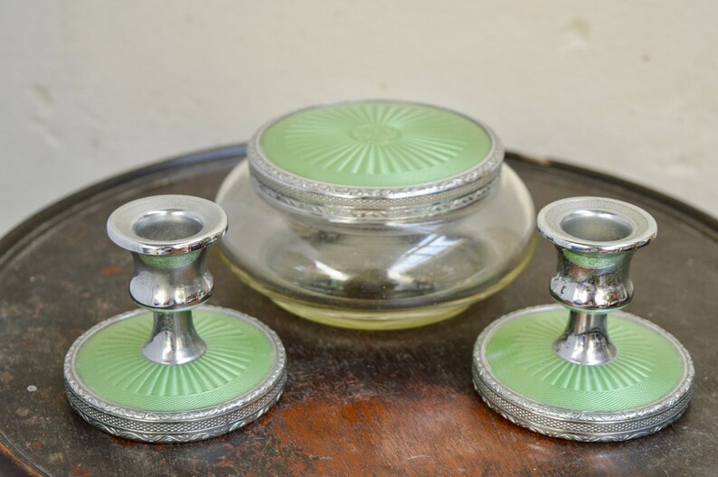 K&ouml;nnte beinhalten: Ein Vintage-Schminktisch-Set mit zwei silbernen Kerzenst&auml;ndern und einem Glasgef&auml;&szlig; mit gr&uuml;nem Deckel. Die Kerzenst&auml;nder haben eine gr&uuml;ne Basis mit einem silbernen Rand.