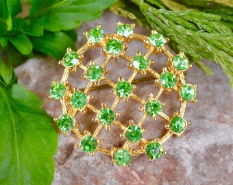 May include: A gold-toned brooch with a circular design featuring a grid pattern of green rhinestones.