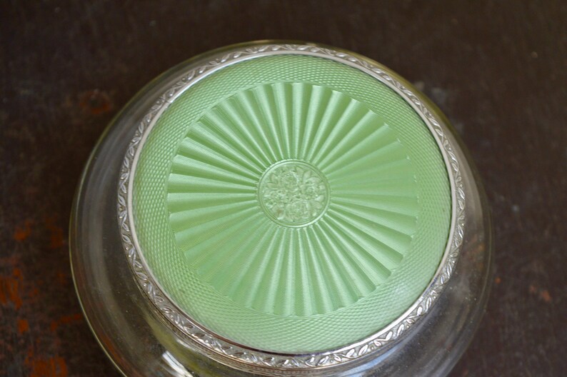 K&ouml;nnte beinhalten: Eine antike Glas-Schmuckschatulle mit silberfarbenem Rand und einem gr&uuml;nen Deckel mit Sonnenstrahlen-Design.