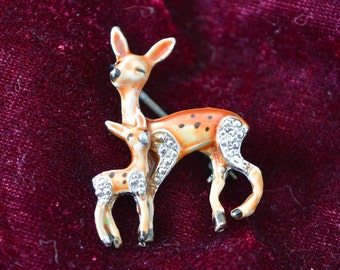 Könnte beinhalten: Eine Vintage-Brosche mit einer Mutter- und einem Jungtier. Die Hirsche sind aus weißem Metall mit braunem und weißem Emaille und haben Strasssteine für die Augen. Die Mutterhirsch steht mit dem Kopf zur Seite gedreht und dem Vorderbein erhoben. Das Jungtier steht neben ihr und schaut zu ihr auf.