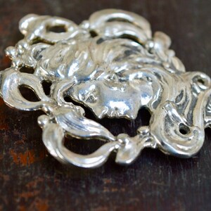 May include: A silver-toned metal pendant featuring a detailed, stylised portrait of a woman's face surrounded by intricate floral designs.