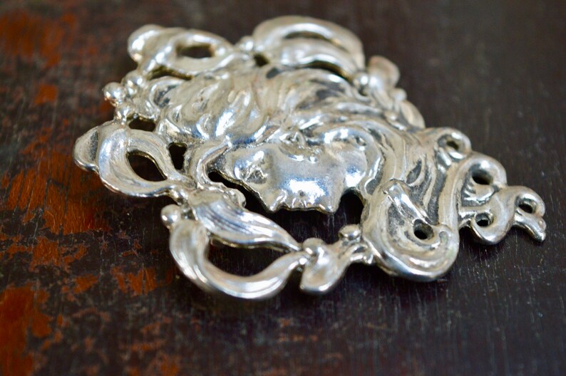May include: A silver-toned metal pendant featuring a detailed, stylised portrait of a woman's face surrounded by intricate floral designs.
