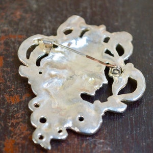 May include: Silver-toned brooch with an intricate design featuring a floral motif. The brooch has a pin and catch fastening on the back.