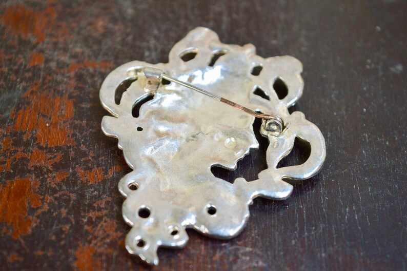 May include: Silver-toned brooch with an intricate design featuring a floral motif. The brooch has a pin and catch fastening on the back.