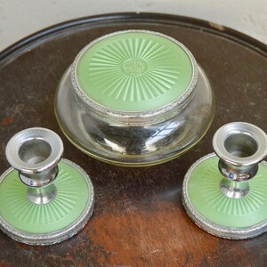 K&ouml;nnte beinhalten: Ein Vintage-Schminktisch-Set mit zwei silbernen Kerzenst&auml;ndern und einem Glasgef&auml;&szlig; mit gr&uuml;nem Deckel. Die Kerzenst&auml;nder und das Gef&auml;&szlig; haben einen dekorativen silbernen Rand und eine gr&uuml;ne Basis.