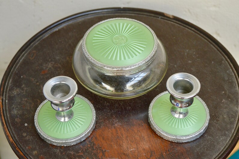 K&ouml;nnte beinhalten: Ein Vintage-Schminktisch-Set mit zwei silbernen Kerzenst&auml;ndern und einem Glasgef&auml;&szlig; mit gr&uuml;nem Deckel. Die Kerzenst&auml;nder und das Gef&auml;&szlig; haben einen dekorativen silbernen Rand und eine gr&uuml;ne Basis.