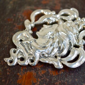 May include: Silver-toned metal ornament featuring a detailed profile of a woman's face with flowing hair, surrounded by intricate floral designs.