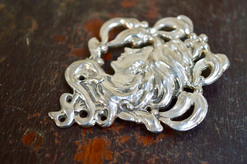 May include: Silver-toned metal ornament featuring a detailed profile of a woman's face with flowing hair, surrounded by intricate floral designs.