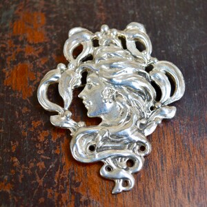 May include: A silver-toned brooch featuring a woman's face in profile, surrounded by intricate floral designs.