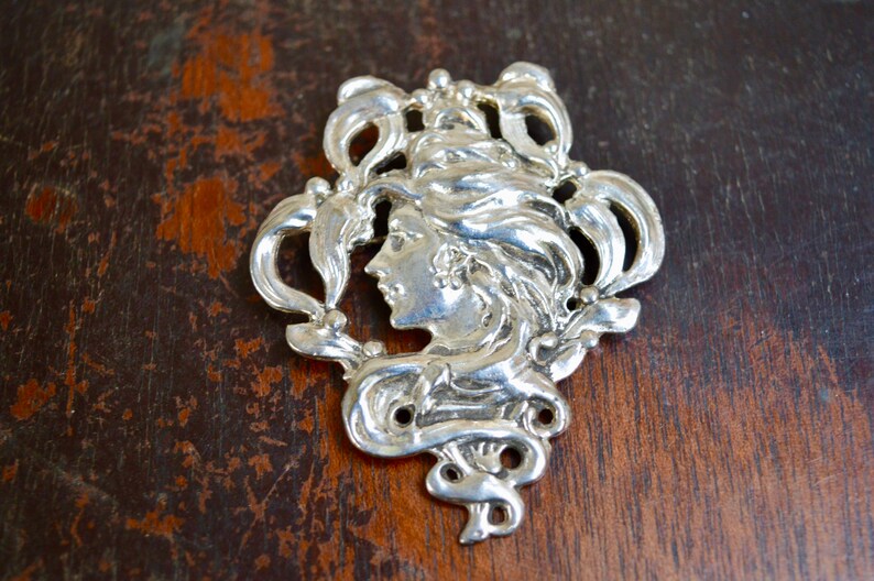 May include: A silver-toned brooch featuring a woman's face in profile, surrounded by intricate floral designs.