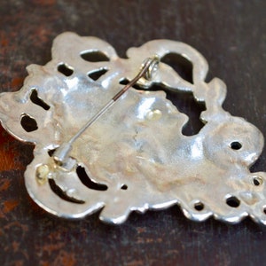 May include: Silver-toned brooch with an intricate, ornate design. The back of the brooch features a pin and catch.