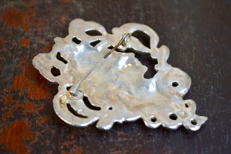 May include: Silver-toned brooch with an intricate, ornate design. The back of the brooch features a pin and catch.