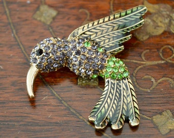 Könnte beinhalten: Eine goldfarbene Kolibri-Brosche mit lila und grünen Strasssteinen. Die Flügel sind mit schwarzem und goldenem Email verziert.