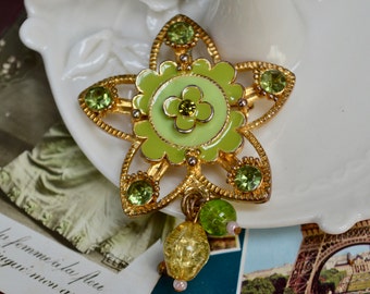 Pode incluir: Um broche em forma de estrela dourado com esmalte verde e strass. O centro da estrela apresenta uma flor verde com um centro amarelo.