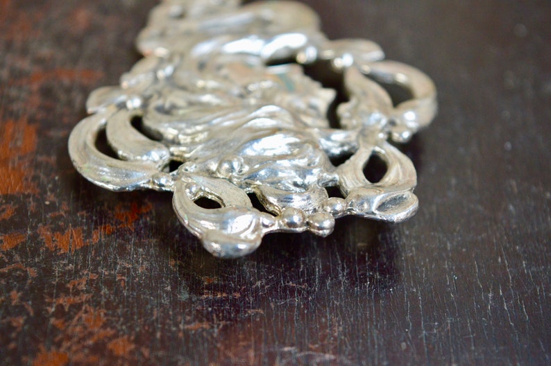 May include: A silver-toned metal ornament with a floral design. The ornament has a detailed, intricate design with a raised centre.