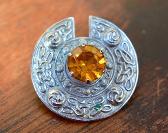 Puede incluir: Un broche de nudo celta de plata con una piedra preciosa de color ámbar en el centro.