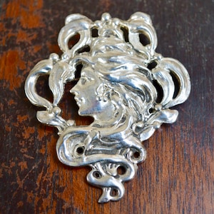 May include: Silver pendant featuring a detailed Art Nouveau design of a woman's face surrounded by stylised floral motifs.