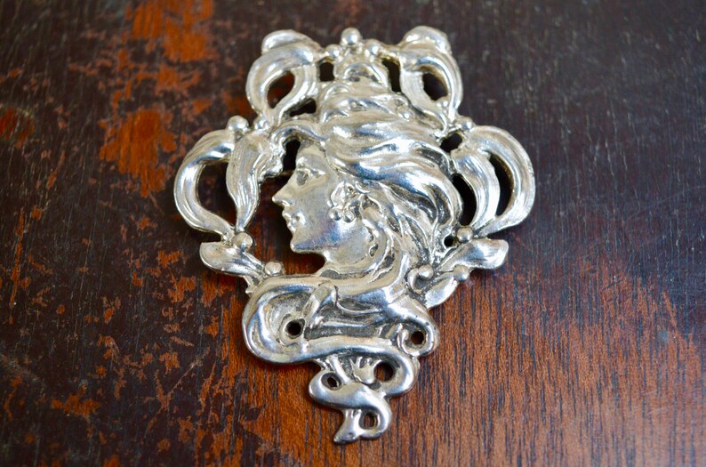 May include: Silver pendant featuring a detailed Art Nouveau design of a woman's face surrounded by stylised floral motifs.