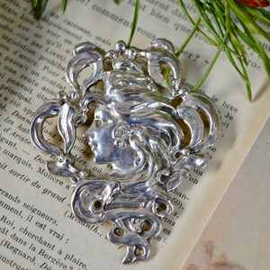 May include: Silver Art Nouveau style brooch featuring a woman's face with flowing hair and intricate floral designs.