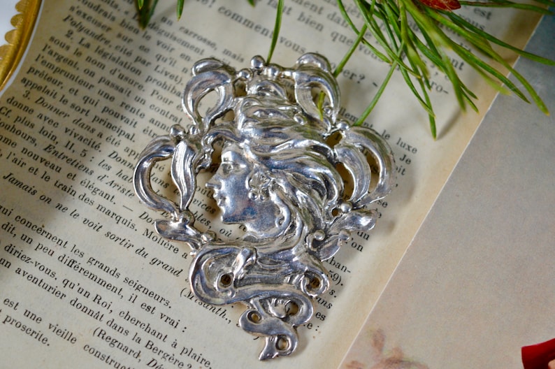 May include: Silver Art Nouveau style brooch featuring a woman's face with flowing hair and intricate floral designs.