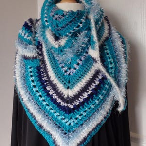 Op de afbeelding: Een handgemaakte gehaakte sjaal in tinten van teal, wit en marineblauw. De driehoekige sjaal heeft een gestructureerd steekpatroon en een pluizige, gefranjerde rand. De sjaal wordt getoond op een houten mannequinhoofd.