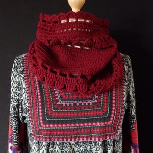 Op de afbeelding: Een bordeauxrode gehaakte cowl sjaal met een geschulpte rand en openwerk ontwerp. De sjaal wordt getoond op een mannequinhoofd, waarbij de textuur en stijl worden getoond. De achtergrond heeft een patroon top met rode en zwarte borduursels.