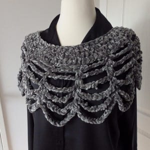 Może przedstawiać: Szary szydełkowy szal o unikalnym wzorze. Szal wykonany jest z miękkiej, pluszowej przędzy i posiada falisty brzeg. Wzór jest otwarty i przewiewny, z szeregiem połączonych pętli. Szal jest prezentowany na czarnej koszuli.