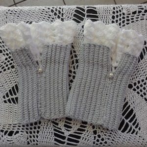 Peut inclure: Une paire de mitaines composée d'un corps en crochet gris côtelé et d'un poignet en crochet blanc à volants. Chaque gant est orné d'une petite perle blanche à la base de l'ouverture du pouce, ajoutant un détail décoratif à l'ensemble.