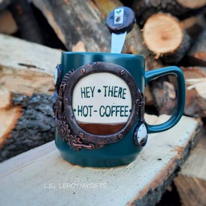 May include: A teal blue ceramic mug with a steampunk design. The mug has a round, white window with the text "Hey There Hot Coffee" inside. The mug has a brown handle and is sitting on a wooden surface.