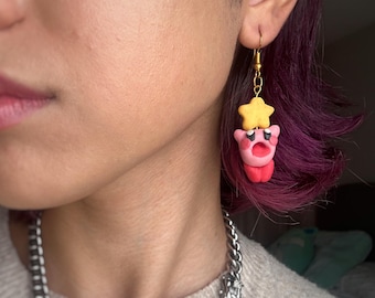 May include: Pink and yellow clay earrings featuring a character with a star on its head. The earrings are hanging from gold hooks.