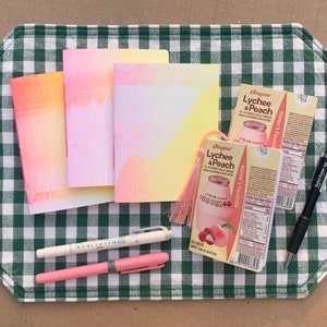 May include: Three small notebooks with watercolor-like designs in shades of pink, orange, and yellow, two black pens, a pink pen, and two boxes of "Binggrae Lychee & Peach Flavored Milk Drink" on a green and white checkered tablecloth.
