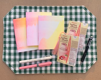 May include: Three small notebooks with watercolor-like designs in shades of pink, orange, and yellow, two black pens, a pink pen, and two boxes of "Binggrae Lychee & Peach Flavored Milk Drink" on a green and white checkered tablecloth.
