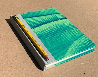 May include: A small, green and blue patterned notebook with a yellow pencil on top. The notebook has a grey spine with the text "DIXON TICONDEROGA" printed on it.