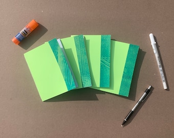 May include: Five small green notebooks with a blue and green patterned design on the spine. The notebooks are arranged in a fan shape on a brown surface. There is a glue stick, a black pen, and a white pen on the surface.